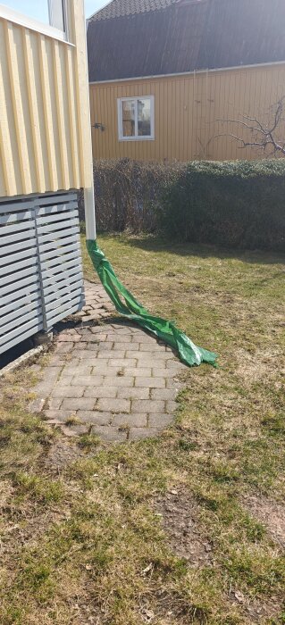 Nedre delen av ett hus med stuprör som mynnar ut i grönt dräneringsrör på en stenlagd gång i en trädgård.