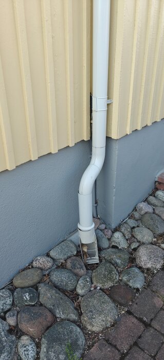 Nedlopp av stuprör som slutar vid marken bredvid en byggnad. Underlaget är täckt av stenar.