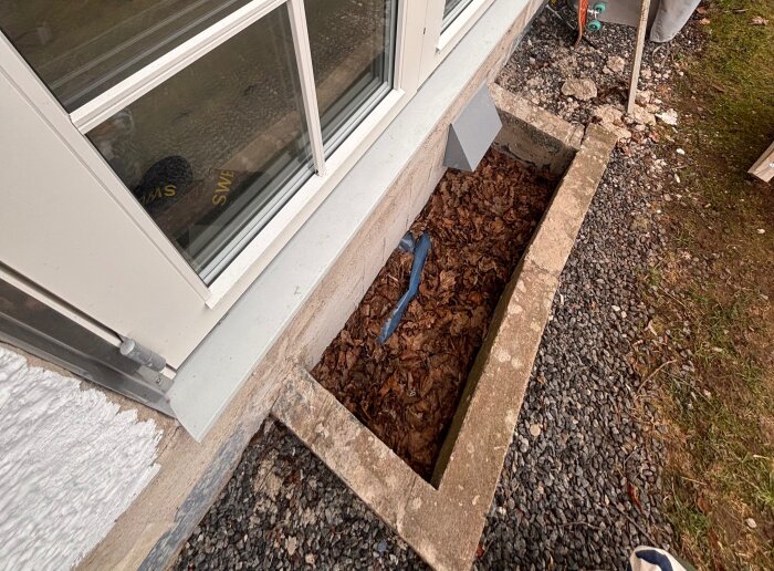 Fönsterkällare med brunn fylld av löv vid husfasad, synlig ventilationstrumma och grusmarkeringar.