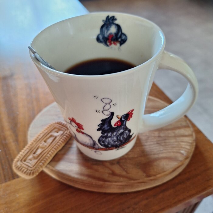 En vit mugg med tuppmotiv fylld med kaffe, stående på ett träunderlägg bredvid en rektangulär kaka.