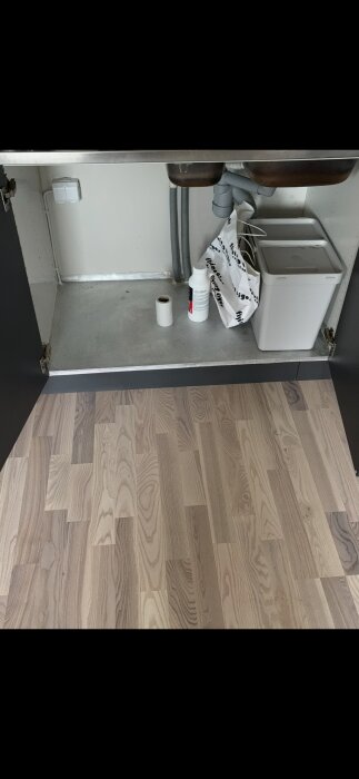 Skåp under diskbänk med rör, rengöringsmedel, plastpåse och avfallskärl på laminatgolv.