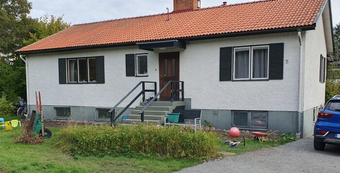 En vit villa med tegelrött tak och mörka fönsterluckor, omgiven av gräsmatta och växter, med en trappa och parkering bredvid.