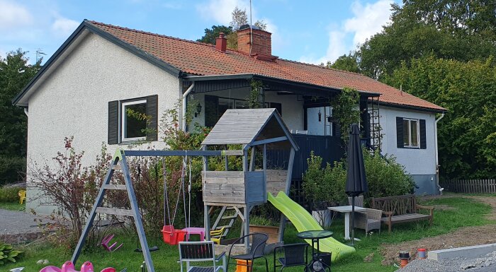 En vit hus med rött tak, en veranda och en lekplats i trädgården med rutschkana och gunga, omgiven av grönska.