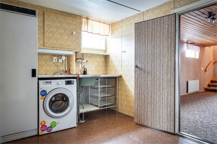 Källare från 1980-talet med tvättstuga, tvättmaskin, diskbänk och öppning mot gillestuga. Retro tapeter och inredning.