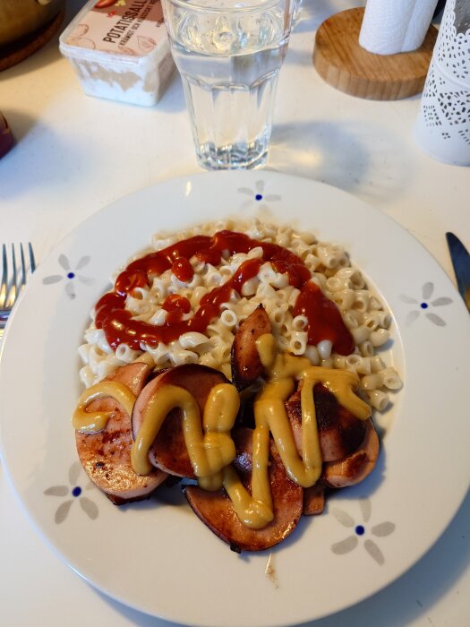 Tallrik med stuvade makaroner med ketchup och stekt falukorv med senap, serverad med ett glas vatten i bakgrunden.