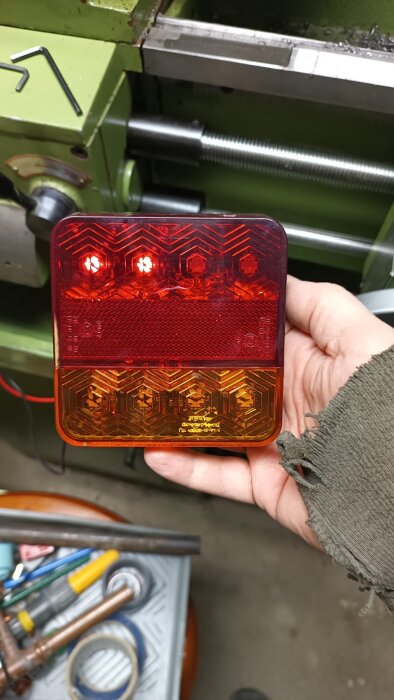 Baklampa visas framför en svarvmaskin, hålls i handen. Röda och gula färger. Besiktning närmar sig.