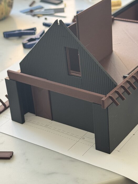 Modell av ett hus i kartong med ett triangulärt tak, väggar och en öppning för ett fönster, placerad på en ritning med verktyg runtom.
