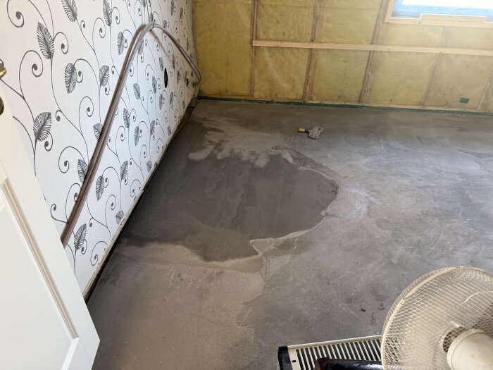 Rum under renovering med nylagt spackel på golvet och tapet med bladmönster. Ett vattenpass ligger på golvet.