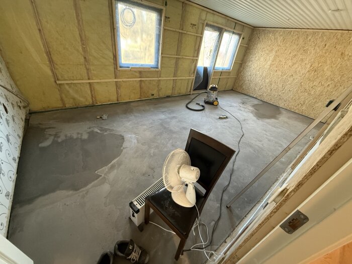 Rum under renovering med ojämnt golv och verktyg, gul isoleringsvägg och plywoodpanel. Fläkt och skor synliga på golvet.