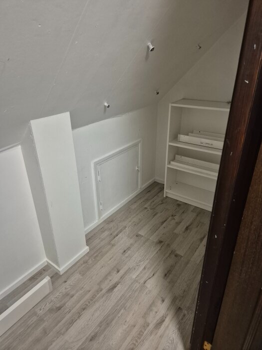 Vitmålat snedtak med installerade spotlights, tomt hörn med vita hyllor och grått trägolv i ett förråd.
