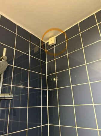 Eldosa i badrum som "danglar" från sladd nära taket, monterad ovanför kakel i duschområde, markerad med orange cirkel.