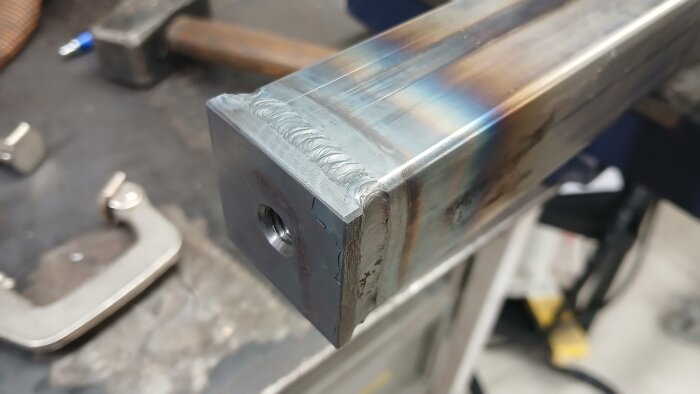 Svetsat metallrör i närbild, arbetsplats synlig i bakgrunden med verktyg och svetsdetaljer.
