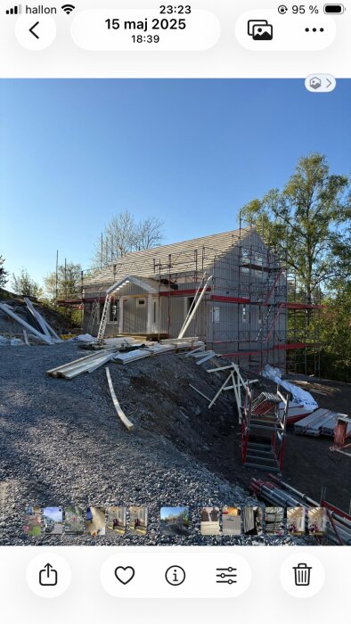 Hus under konstruktion med byggnadsställningar och material i grusig omgivning, klar himmel och träd i bakgrunden.
