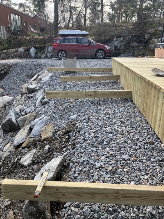 Trädäck under uppbyggnad med träreglar på grus. I bakgrunden står en röd bil parkerad framför en stenmur och en tegelbyggnad.