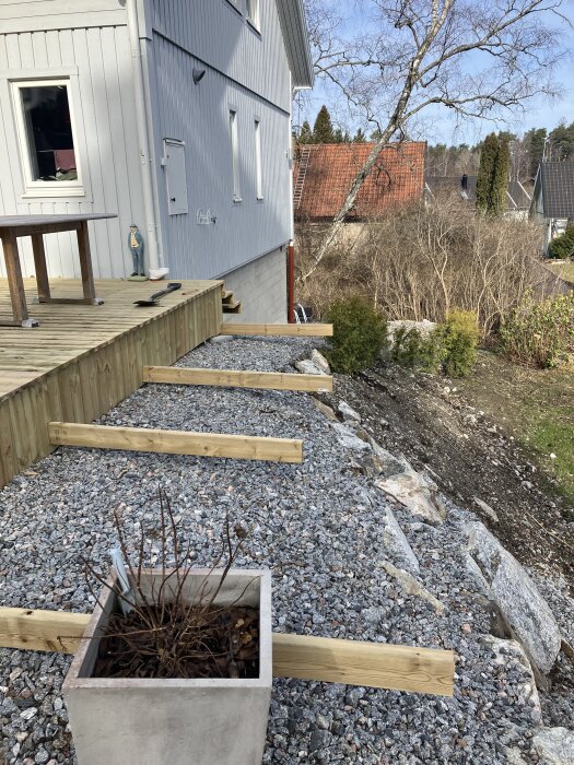 Träbjälkar på grusmark vid sidan av ett hus, med veranda och planteringskruka i förgrunden. Träd och buskar i bakgrunden.