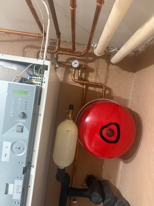 Rörsystem med manometer och expansionskärl, kopparkablar och plastledningar i ett tekniskt rum. Ett rött kärl har en markerad svart cirkel.