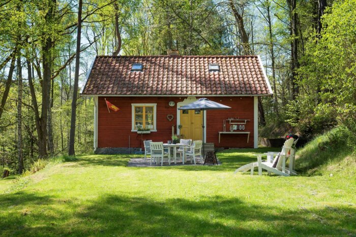 Rött trähus med tegeltak i grönskande omgivning, trädgårdsbord med parasoll framför, solstolar på gräsmattan, mysig skogsmiljö.