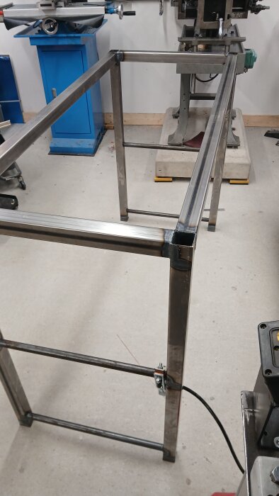 Metallram för arbetsbord under konstruktion, med fokus på svetsade hörn och stålben, belägen i en verkstadsmiljö med verktygsutrustning.