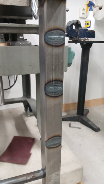 Närbild av svetsfogar på metallben till ett arbetsbord, där flera svetsar syns på rad och verktyg finns i bakgrunden.