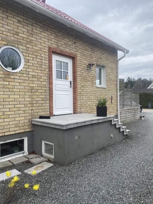 Tegelhus med vit dörr, stentrappa utan räcke och plattform utan tak. Grusgång med blomkruka bredvid trappan. Hund syns i bakgrunden.