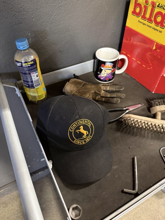 Arbetsbänk med verktyg och tillbehör, inklusive svart keps med gul logotyp, arbetsvantar, kaffekopp, trådborste, skiftnyckel och dryckesflaska.