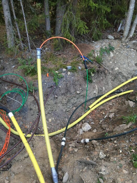 Skyddsslangar och oterminerade fiberkablar vid en grävd tomt omgiven av skog, förberett för fiberinstallation i ett hus.