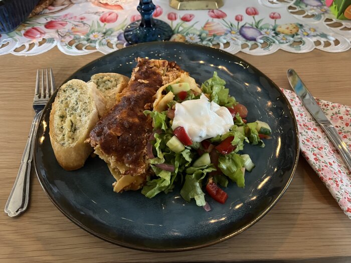 Tacolasagne med riven ost och sallad toppad med vitlökssås på en tallrik, serverad med vitlöksbröd på ett dukat bord.