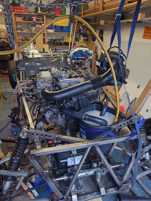 Bilmotor i garage monterad i ram med slangar och vajrar, omgiven av verktyg och hyllor fulla med verkstadsutrustning.