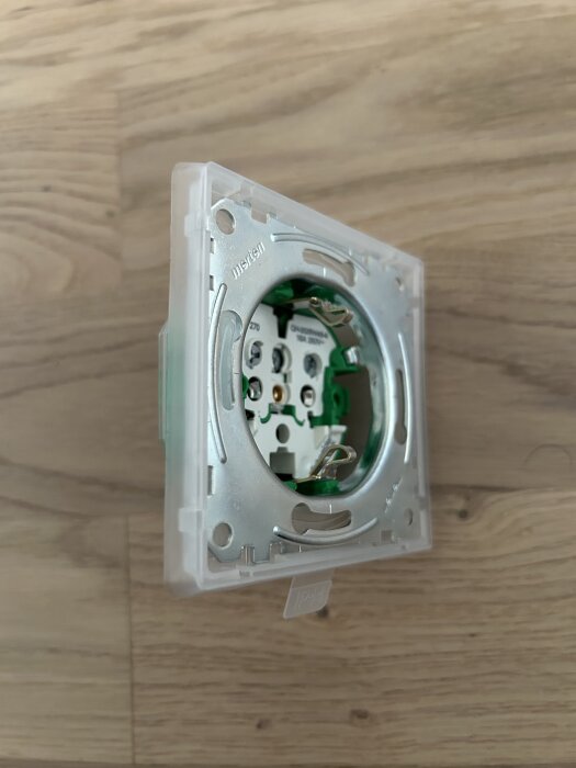 Elektrisk vägguttag utan kåpa, sedd från sidan, monterad på trägolv, visar metall- och plastkomponenter samt skruvkopplingar.