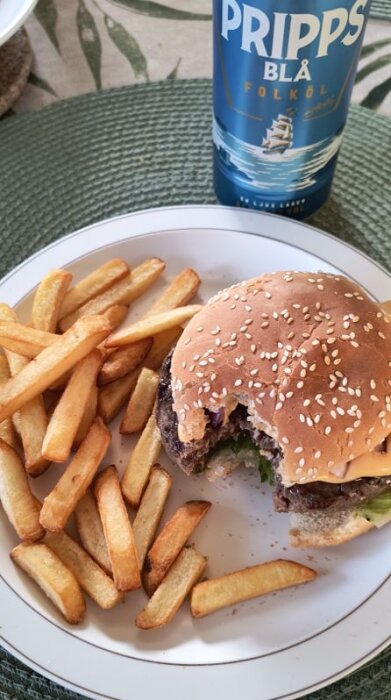 Hamburgare med bacon och ost på tallrik bredvid pommes frites, Pripps Blå folköl i bakgrunden.