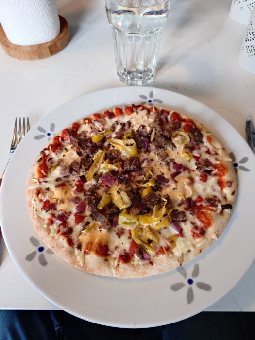 Fryspizza med topping av kött, lök och paprika på tallrik, bredvid ett glas vatten på ett bord.