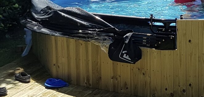 Byggprojekt av träpanel runt pool med pågående regelfäste till metallram, synlig poolvattenyta och trätrall med skor och klädsel.