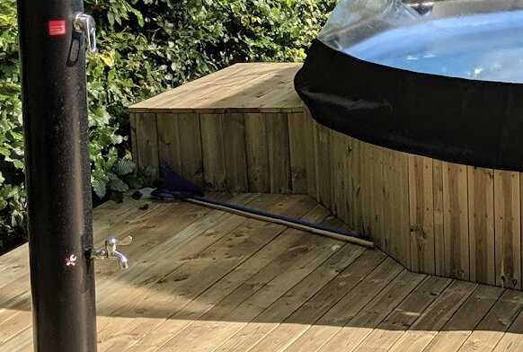 Träaltan med poolinsats och duschstång i ett trädgårdsområde. Plattform byggd för att dölja slangkopplingar, synliga verktyg på marken.