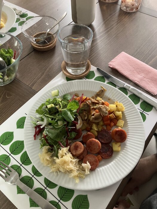 Tallrik med pyttipanna-variant: potatis, morot, svamp, lök, Kolbasz, sallad och surkÄl. Placerad pÄ bord med glas vatten och servett.
