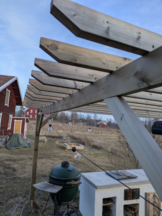 Pergola med träbalkar och dekorativa lampor i en trädgård, med ett rött hus och öppen mark i bakgrunden.
