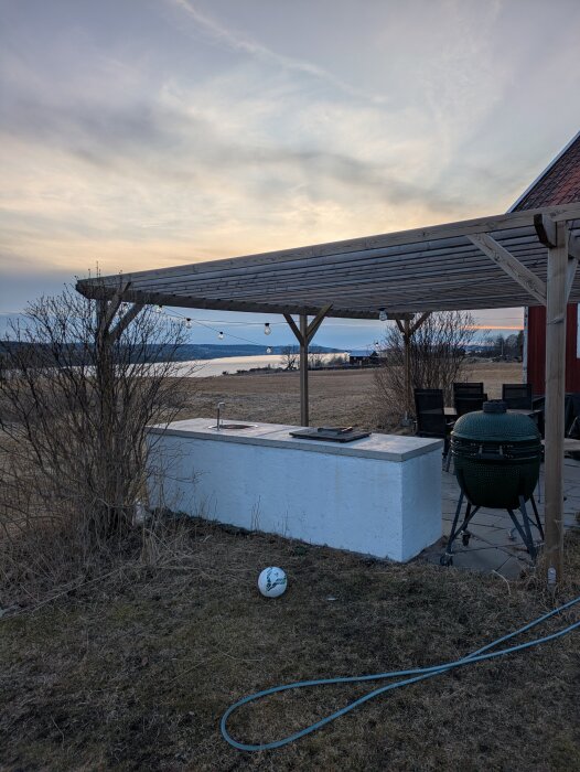 Pergola med takbjälkar över uteplats, vita tegelblock och en stor grill i trädgården, eftermiddagsslöja med vatten i bakgrunden.