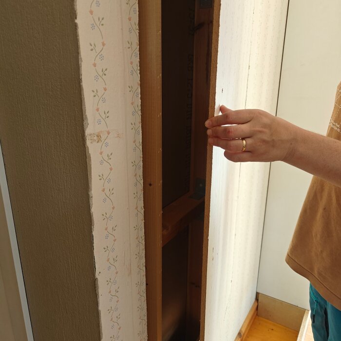 Hand som inspekterar vägg med borttagen panel, visar bakomliggande struktur och äldre tapet i en hall under renovering.