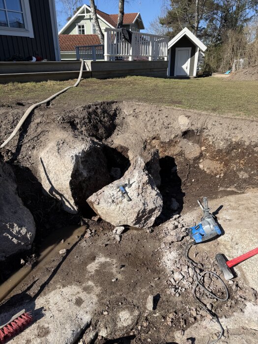 Grävt hål i marken med stora stenar utspridda, en borrmaskin, slägga och markeringsverktyg syns. En veranda och liten byggnad finns i bakgrunden.