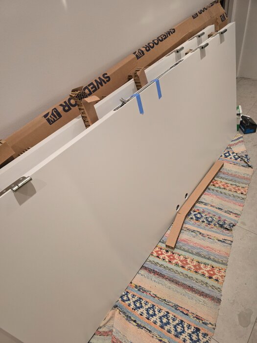 En vit dörr från Swedoor ligger på en mönstrad matta, omgiven av förpackningsmaterial och med gångjärn synliga på ena sidan.