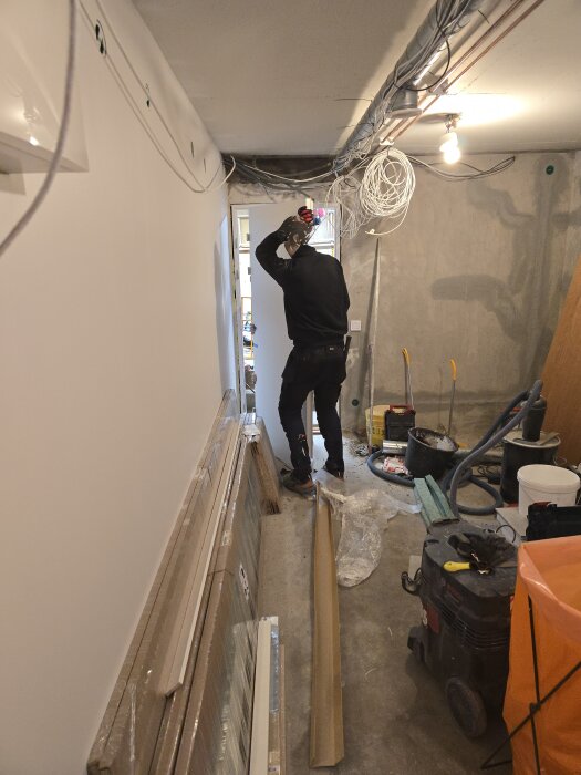 Person monterar dörrkarm i smal hall med byggmaterial och verktyg runt om. Spackelämbar och sladdar synliga på betongvägg.