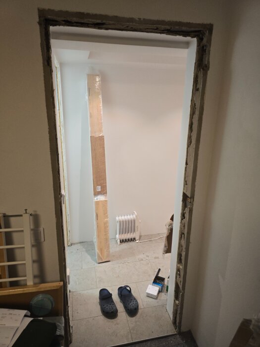 Öppning för en dörrinstallation i ett rum under renovering, med dörrkarmar, två blå tofflor, en elektrisk radiator och byggmaterial synliga.
