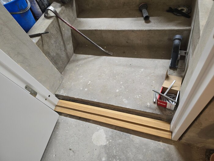 Tröskel av trä installeras mellan en källardörr och ett betonggolv med byggverktyg och material synliga i bakgrunden.