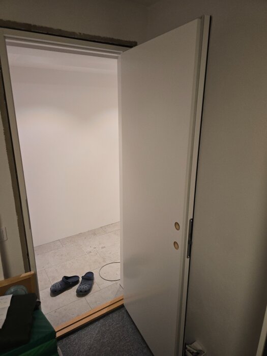 Vit innerdörr med öppen position, visar övergång mellan två rum. På golvet finns ett par svarta tofflor och en sladd som ligger löst.