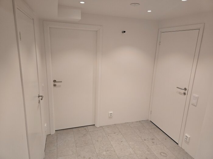 Vitmålad hall med tre dörrar i olika riktningar. Klinkergolv och belysning i taket. Rummet är ljust och stilrent inrett.