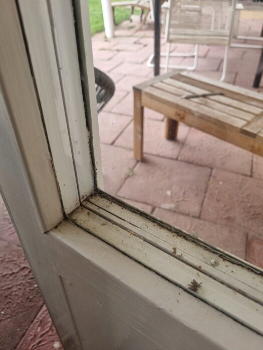 Närbild av en dörrkarm med glas, smuts i hörnet; en uteplats med stenplattor och utemöbler syns genom glaset.