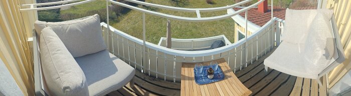 Balkongmöblering med två beige fåtöljer och ett litet träbord, placerade på en träterrass med utsikt över en gräsmatta.