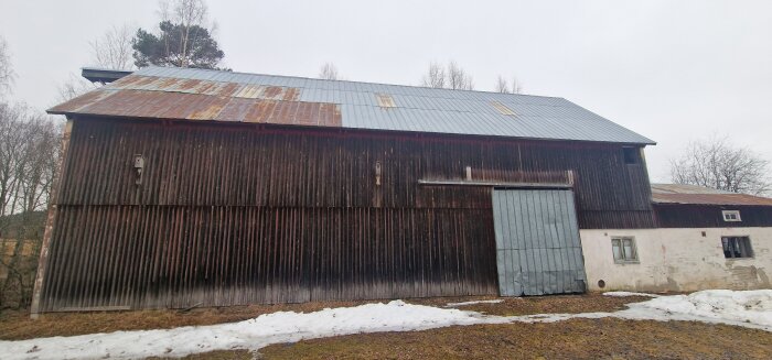 Lada med rostigt plåttak, mörka brädväggar och en stor skjutdörr. Del av vit murad sektion med små fönster syns också. Snöhögar framför ladan.