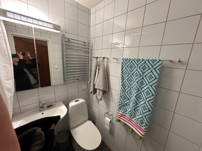 Badrum med vitt kakel från 2003, handfat och toalett. En färgglad handduk på väggen och ett litet hål syns i väggen.