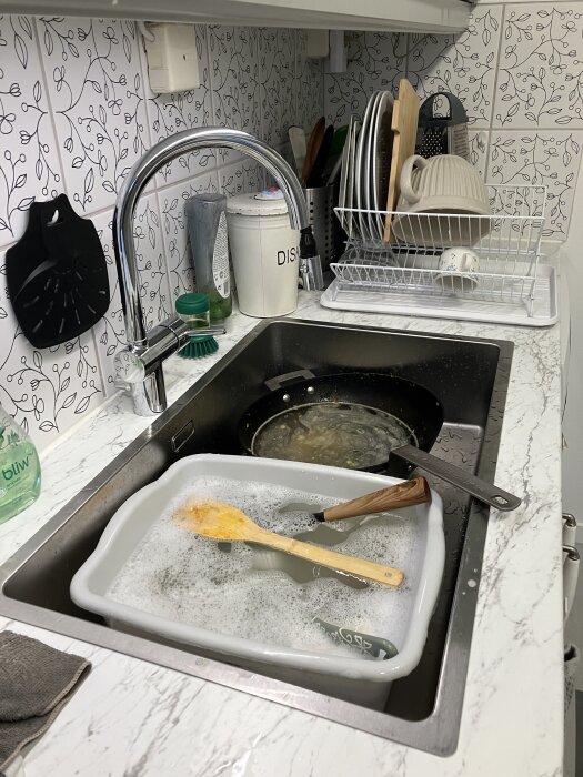 Kök med diskho, diskställ och diskredskap på bänk. Diskborste och skummande vatten syns i balja i vasken.