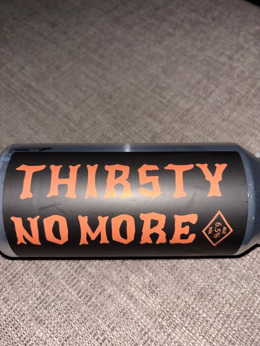 En ölburk med etiketten "Thirsty No More" och alkoholhalt 6,5% vol, liggande på en yta.